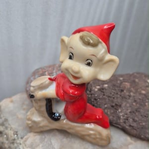 May include: A ceramic figurine of a small elf with large ears, wearing a bright red outfit and a matching pointed hat. The elf is depicted in a kneeling position, resting its hands on a textured, light brown log-shaped base with a dark accent.