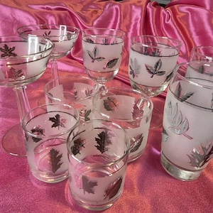 Op de afbeelding: Een verzameling heldere glazen drinkwaren met matte bladeren. De set bevat champagneglazen met steel en diverse tumblers, allemaal met zwarte en zilveren bladerpatronen. De glazen zijn gerangschikt op een roze satijnen oppervlak.