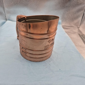 May include: A copper-colored sifter with a wooden handle. The sifter has a cylindrical shape with horizontal ridges and the text "Foley Sift-Chine" on the side. The sifter is designed for sifting flour or other dry ingredients.