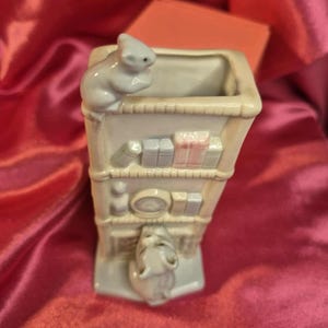 May include: A light-colored ceramic figurine of a building with a mouse on top and another at the bottom. The building has shelves with small books and a clock. The figurine is on a pink background.