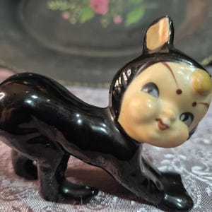 Peut inclure: Figurine vintage en céramique représentant un chat noir en position rampante. Le chat a un visage crème avec des yeux bleus, des lèvres rouges et une tache jaune sur le front. L'objet est un objet de collection.