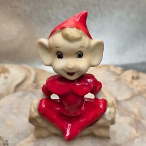 Könnte beinhalten: Eine kleine Keramikfigur eines Elfen mit großen Ohren, der eine leuchtend rote Zipfelmütze und ein passendes rotes Outfit trägt. Der Elf hat einen hellen Teint, dunkle Augen und sitzt mit gekreuzten Beinen, versehen mit einer glänzenden Oberfläche.