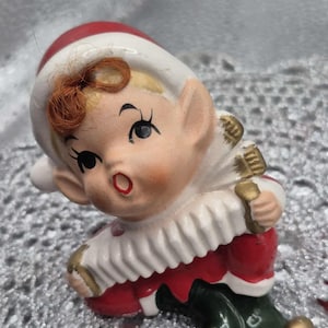 May include: A ceramic Christmas elf figurine, with a red and white hat, a white collar, and a red outfit. The elf has a surprised expression and is holding a golden accordion. The figurine has green shoes with golden accents.