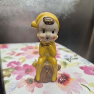 Könnte beinhalten: Eine kleine Keramikfigur eines Charakters in einem leuchtend gelben Kapuzenoutfit, der auf einem braunen Baumstumpf sitzt. Die Figur hat ein blasses Gesicht, rosige Wangen, dunkelbraunes Haar unter der Kapuze und große Augen mit langen Wimpern.