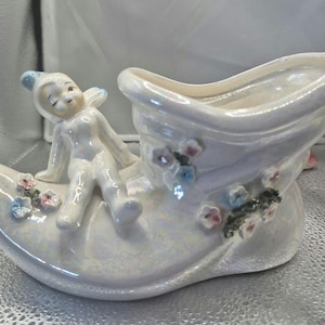 Könnte beinhalten: Ein weißer Keramik-Schuh-Pflanztopf mit einer fantasievollen Feenfigur, die darauf sitzt. Der Schuh ist mit rosa und blauen Blumen verziert.