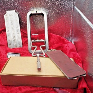 以下が含まれることがあります： 段ボール箱に入ったヴィンテージの理髪用具のセット。道具には、金属製の剃刀、金属製のハンドル、金属製の剃刀の刃が含まれています。道具は赤いベルベットの表面に置かれています。