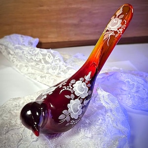 Puede incluir: Figurita decorativa de pájaro de cristal en tonos rojos, naranjas y marrones. El pájaro está adornado con diseños florales blancos. La figura descansa sobre una tela de encaje blanco.