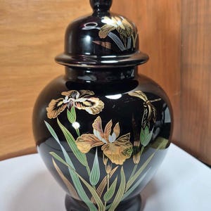May include: A black ceramic jar with a lid, adorned with gold and white iris flower designs. The jar has a rounded body and a pedestal base. The lid has a small knob on top.