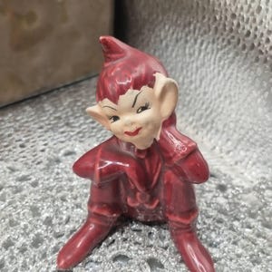 Puede incluir: Una pequeña figura de cerámica de un elfo sentado con un traje rojo oscuro y un gorro puntiagudo a juego. El elfo tiene la cara pálida, orejas puntiagudas y rasgos faciales pintados que incluyen cejas oscuras, ojos almendrados y labios rojos.