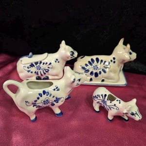 Może przedstawiać: Zestaw ceramicznych naczyń do serwowania w kształcie krowy. Zestaw zawiera dzbanuszek do śmietanki, naczynie z pokrywką, maselniczkę i mały element dekoracyjny. Każdy element jest biały z niebieskimi wzorami kwiatowymi.
