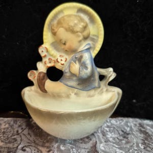 May include: A vintage porcelain figurine of a child seated in a bowl. The child wears a blue jacket and has a yellow halo. Small red and white flowers are on the side. The figurine is a cream colour with brown accents.