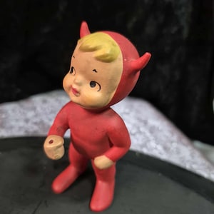 May include: A vintage figurine of a small devil character. The figure is dressed in a red jumpsuit and a matching hood with small horns. The face is light-colored with yellow hair, and the figure is holding a small, beige object.