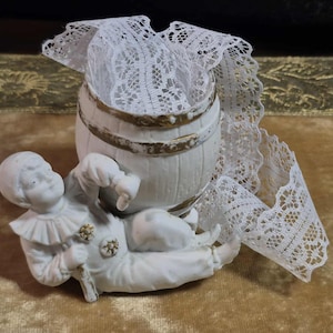 May include: A white porcelain figurine of a clown lying on its side, holding a small cane. A small white barrel with gold trim is behind the clown, with white lace spilling out. The figurine is on a gold velvet surface.