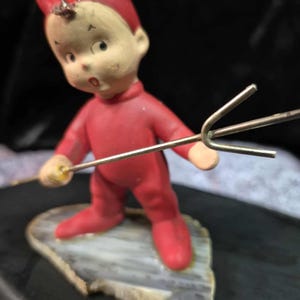 May include: A vintage figurine of a red devil with a surprised expression, wearing a red jumpsuit and horns. The devil holds a small trident and stands on a stone-like base. The figurine has a painted face with a light skin tone.