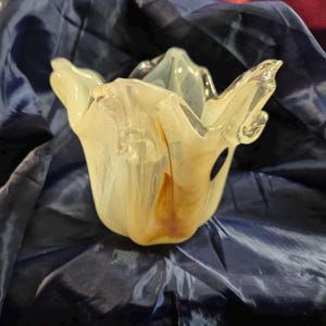 May include: A decorative glass bowl with a unique, organic shape. The bowl features a creamy white and amber color palette, with a flowing, petal-like design. The glass has a glossy finish and is set against a dark blue background.