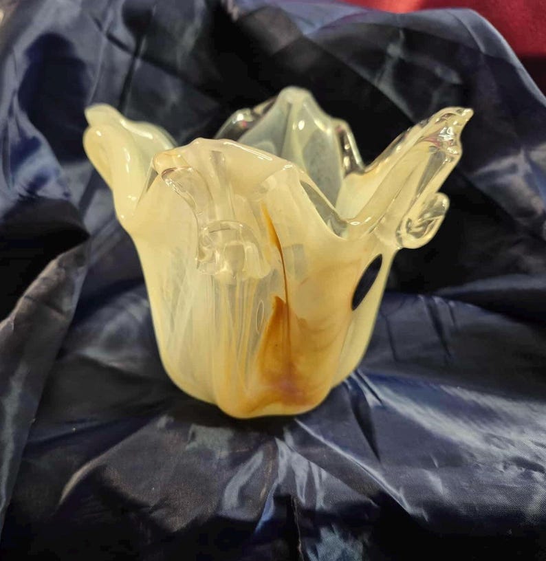 May include: A decorative glass bowl with a unique, organic shape. The bowl features a creamy white and amber color palette, with a flowing, petal-like design. The glass has a glossy finish and is set against a dark blue background.