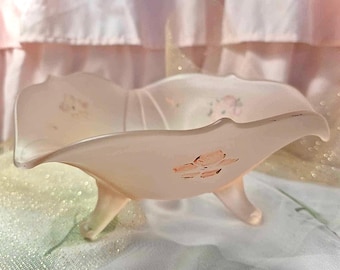 May include: A pale pink, footed dish with a scalloped rim. The dish has a floral design in shades of pink and peach. The dish is made of glass and has three small feet.
