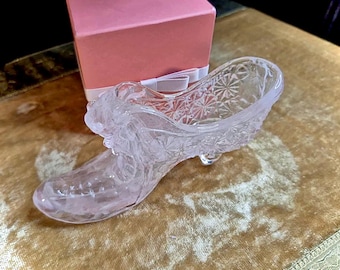 May include: A decorative clear glass slipper with detailed floral and starburst patterns. The shoe is on a gold-toned surface next to a pink gift box with a white ribbon. The shoe has a small heel.