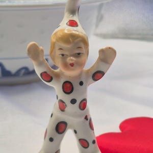 May include: A small, ceramic figurine of a child-like figure in a white jumpsuit with red and black polka dots. The figure has a pointed hat with red accents and is posed with arms raised. A red heart-shaped object is in the background.