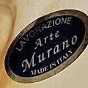 May include: A close-up of an oval sticker with the text "LAVORAZIONE Arte Murano MADE IN ITALY" in white lettering on a dark blue background. The sticker has a gold border and is slightly angled.