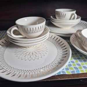 May include: A set of vintage white dishware with gold and black geometric designs. The set includes plates, bowls, and teacups with matching saucers. The teacups have a decorative gold pattern around the rim.