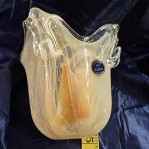 May include: A clear glass vase with a unique, organic shape. The vase has a creamy white and peach color scheme, with a label that reads "Lavorazione Arte Murano". The vase is approximately 5 inches tall.