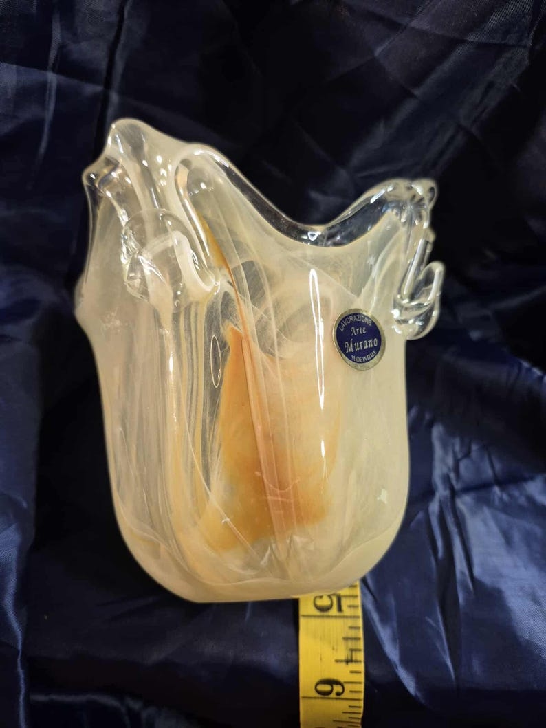 May include: A clear glass vase with a unique, organic shape. The vase has a creamy white and peach color scheme, with a label that reads "Lavorazione Arte Murano". The vase is approximately 5 inches tall.