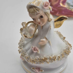 May include: A porcelain figurine of a young girl in a white dress, holding a basket of flowers. The figurine has gold accents on the wings, dress trim, and hat. Pink roses adorn the hat and base. The figurine is approximately 4 inches tall.
