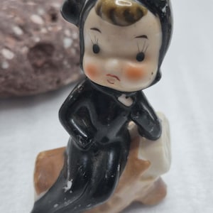 May include: A small ceramic figurine of a child wearing a black hooded outfit and sitting on a brown log. The figure has a pale face with rosy cheeks, dark painted eyes, and a small tuft of brown hair visible under the hood. The finish is glossy.