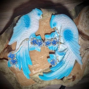 May include: Two decorative bird sculptures in shades of blue and white, perched on branches with blue and gold floral accents. The birds face each other, creating a symmetrical design. The background is a muted gold color.