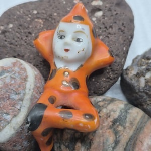 May include: A small ceramic figurine of a jester wearing an orange suit with black polka dots. The figure has a white face with painted features, a pointed hat, and is depicted in a seated position with arms resting behind the head.