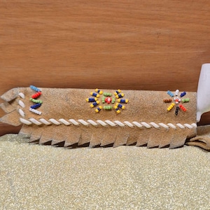 May include: A tan leather pipe case with colorful beaded floral designs and a white wooden pipe. The case has a fringed edge and a braided white cord detail. The beaded designs include red, yellow, blue, and green beads.