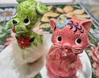 Puede incluir: Una figura de perro de cerámica verde con una flor roja y una figura de gato de cerámica rosa con una flor azul. Ambas figuras de animales tienen un borde de piel sintética blanca en la base y están sobre una superficie reflectante.