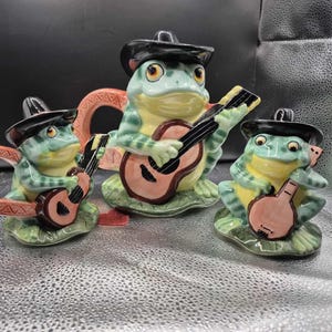 Puede incluir: Un juego de té de cerámica con una banda de ranas. El juego incluye una tetera y dos tazas, cada una con la forma de una rana tocando la guitarra y con un sombrero negro. Las ranas son verdes, amarillas y marrones, con una guitarra rosa.