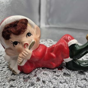 Peut inclure: Figurine de lutin de Noël en céramique en position couchée. Le lutin porte un costume rouge avec des bordures blanches, des chaussures vertes avec des accents dorés et un chapeau blanc. Le lutin a un visage peint avec des cheveux bruns.