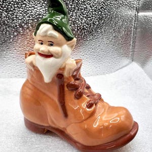Puede incluir: Una figurita de gnomo de cerámica con barba blanca y sombrero verde, asomándose de una bota marrón.