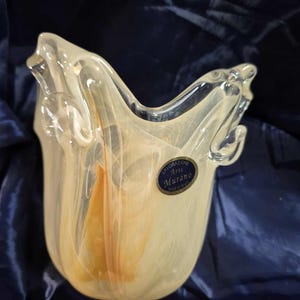 May include: A clear glass vase with a unique shape and a label that reads "Lavorazione Arte Murano Made in Italy." The vase has a creamy yellow and orange color scheme and a textured appearance.
