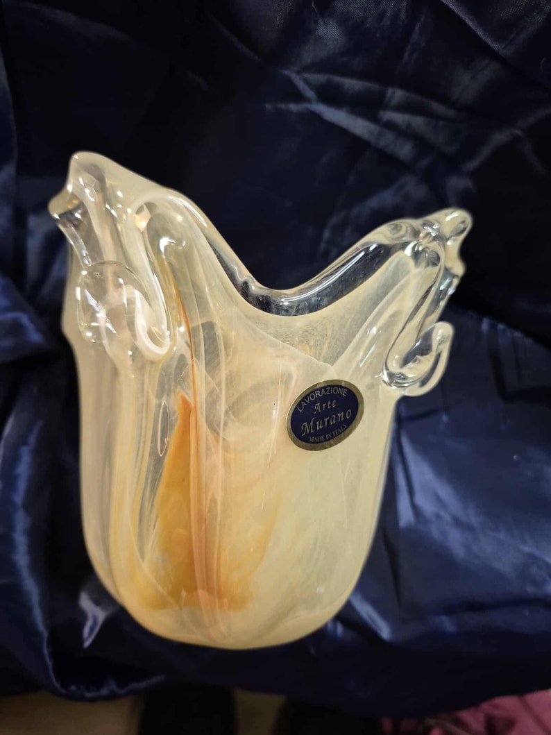 May include: A clear glass vase with a unique shape and a label that reads "Lavorazione Arte Murano Made in Italy." The vase has a creamy yellow and orange color scheme and a textured appearance.