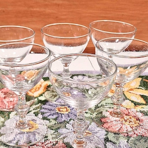 May include: Six clear, stemmed champagne glasses with rounded bowls. The glasses sit on a floral patterned runner with pink, purple, and yellow flowers. The stems have a decorative design. The background is a wooden surface.