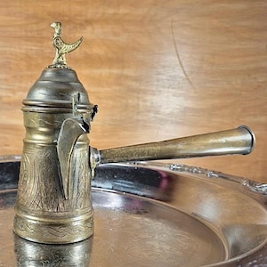 May include: Antique brass coffee pot with a bird-shaped finial. The pot features intricate etched designs and a long, curved spout. It sits on a silver-colored tray, with a wooden background. A vintage kitchenware item.