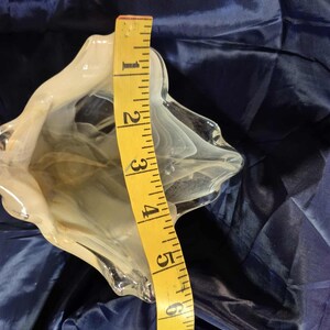 May include: A clear glass decorative bowl with a white swirl design. The bowl is approximately 8 inches tall, as measured by a yellow measuring tape. The bowl has a unique, organic shape and sits on a blue surface.