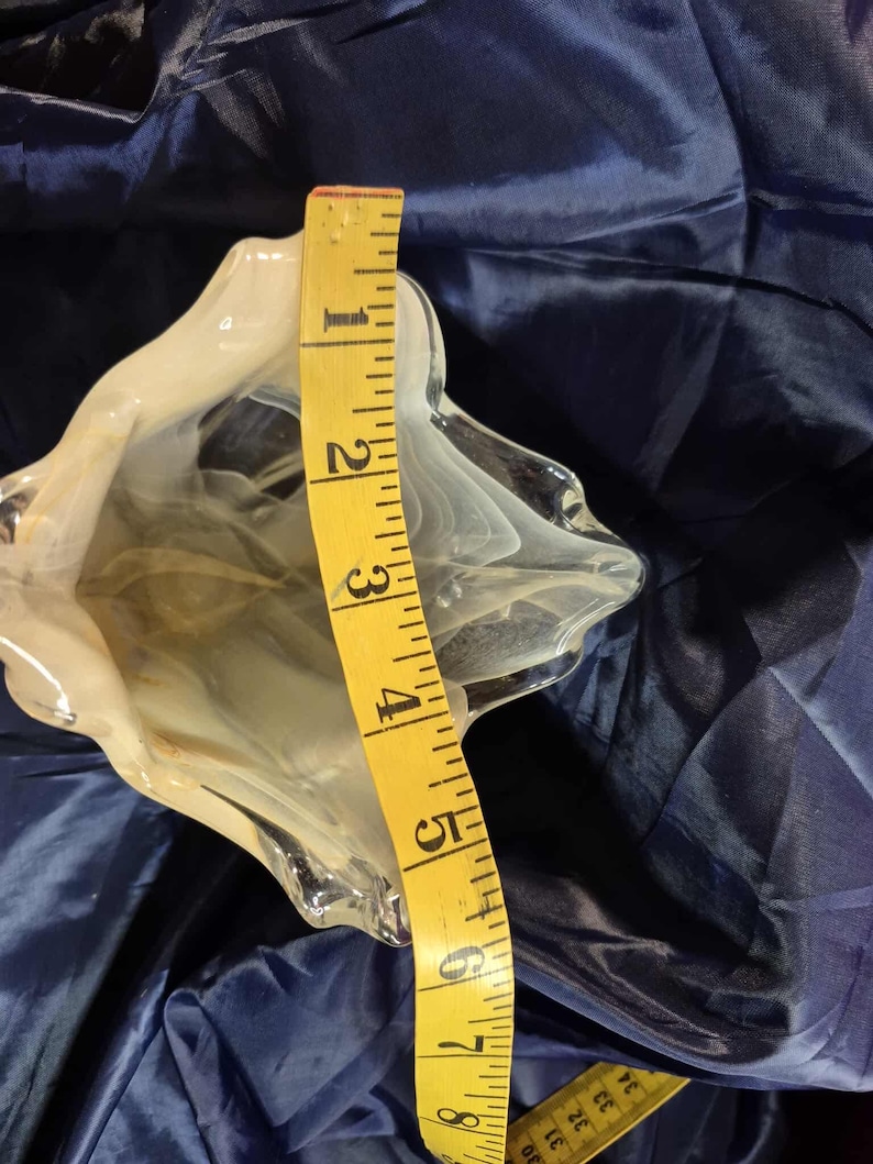 May include: A clear glass decorative bowl with a white swirl design. The bowl is approximately 8 inches tall, as measured by a yellow measuring tape. The bowl has a unique, organic shape and sits on a blue surface.