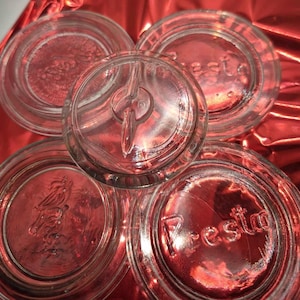 Op de afbeelding: Een verzameling heldere glazen potdeksels en een deksel met een handvat. De deksels hebben reliëfontwerpen en het woord "Presto". De deksels zijn gerangschikt op een rode, reflecterende ondergrond.