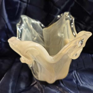 May include: A decorative glass bowl with a unique, organic shape. The bowl is a creamy white color with swirling, translucent patterns. The edges of the bowl have a ruffled, petal-like design, adding to its artistic appeal. The bowl is likely used for decorative purposes.