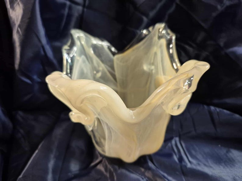 May include: A decorative glass bowl with a unique, organic shape. The bowl is a creamy white color with swirling, translucent patterns. The edges of the bowl have a ruffled, petal-like design, adding to its artistic appeal. The bowl is likely used for decorative purposes.