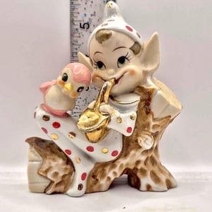 Puede incluir: Figura de cerámica vintage de un elfo sentado con un pájaro. El elfo viste un atuendo blanco con lunares rojos, un chaleco marrón y un sombrero blanco con lunares rojos. El elfo sostiene un objeto dorado. El pájaro es rosa y blanco.