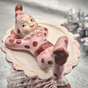 May include: A vintage ceramic figurine of a clown in a pink polka-dot outfit, reclining on a white scalloped plate. The clown has a smiling face and is resting on a decorative base with red and white stripes.