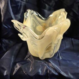 May include: A decorative glass bowl with a unique, organic shape. The bowl is a pale yellow color with white marbling, and it sits on a dark blue surface. The bowl's edges are curved and flared, creating a petal-like appearance.