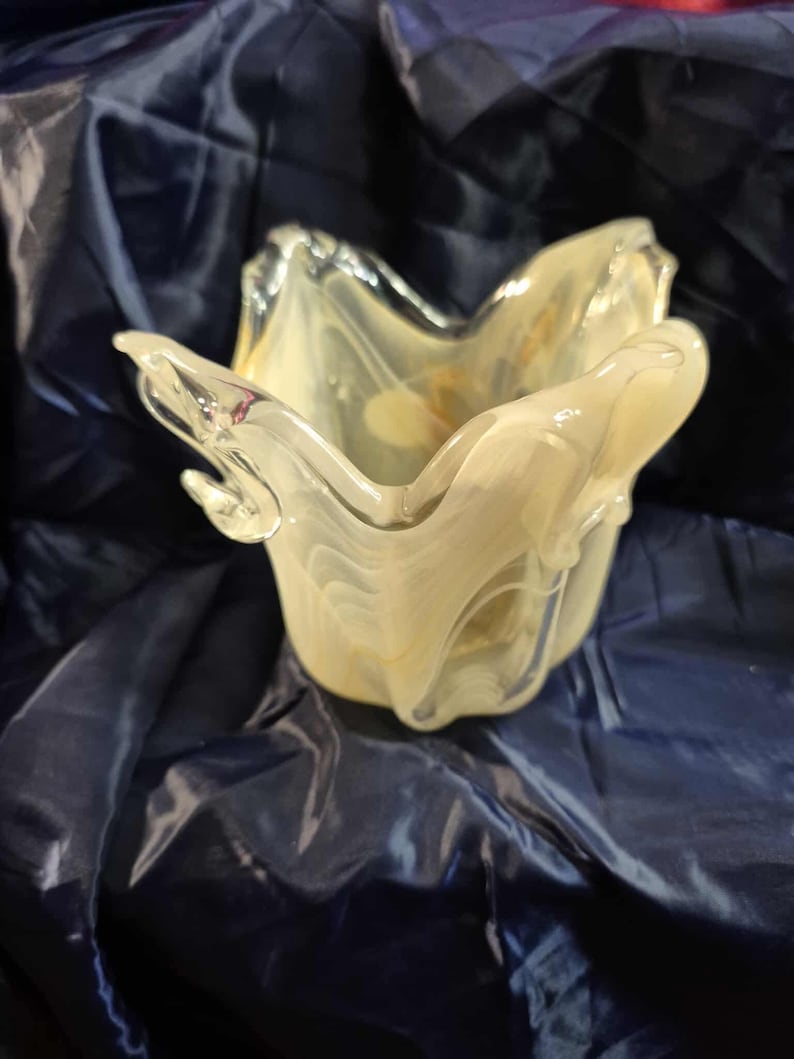May include: A decorative glass bowl with a unique, organic shape. The bowl is a pale yellow color with white marbling, and it sits on a dark blue surface. The bowl's edges are curved and flared, creating a petal-like appearance.
