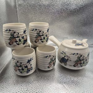 Puede incluir: Un juego de té de cerámica blanca con una tetera y cuatro tazas. Cada pieza presenta una ilustración de dibujos animados de figuras con ropa colorida, con detalles florales azules. Las tazas tienen un diseño acanalado.